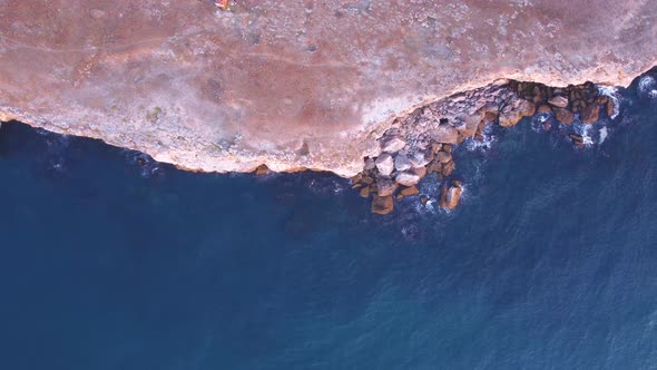 Top down aerial view of waves splash against rocky seashore, background. Flight over high cliffs of