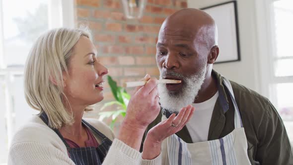 Mixed race senior couple wearing aprons tasting food while cooking in the kitchen at home