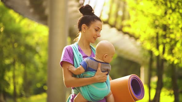 Sporty Woman Carrying Baby in Sling and Walking To Yoga Class in Park