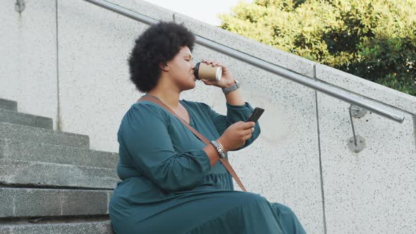 Happy plus size biracial woman using smartphone and drinking coffee