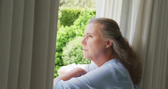 Senior caucasian woman wearing blue shirt and looking through window in living room