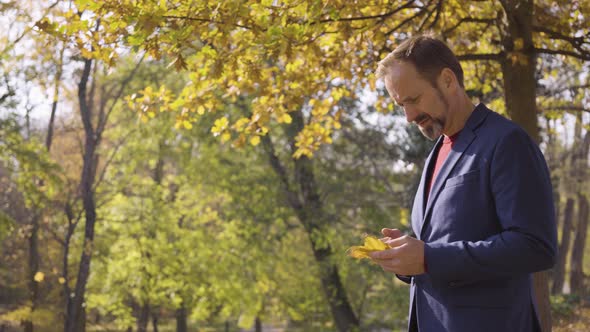 A Middleaged Caucasian Man Examines a Leaf He Holds and Looks Around in Thought in a Park in Fall