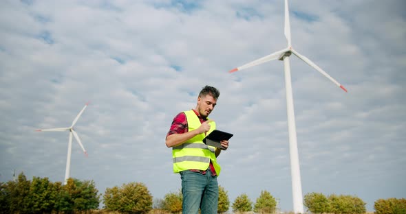 The Engineer Work Next to the Windmill Turbines with the Tablet in His Hand