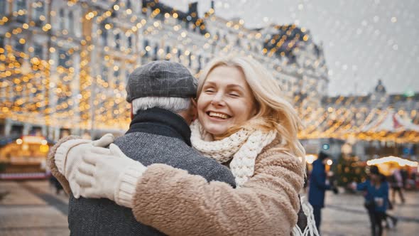 Happy Senior Blonde Woman Running to Man Hugging Him Enjoying Christmas Meeting on City Square