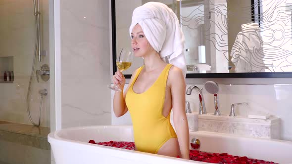 Sexy Woman in Swimsuit with White Wine Relax in Bathtub with Flowers Petals