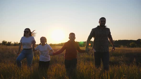 Mother and Father with Two Kids Jog Through Grass Field Holding Hands of Each Other at Sunset