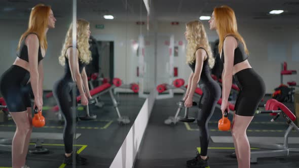 Slim Redhead Sportswoman Talking Lifting Weights with Blond Friend in Gym Looking at Reflection in