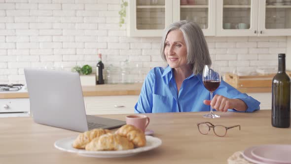 Happy Senior Lady Drinking Wine Video Calling Friend on Laptop at Home