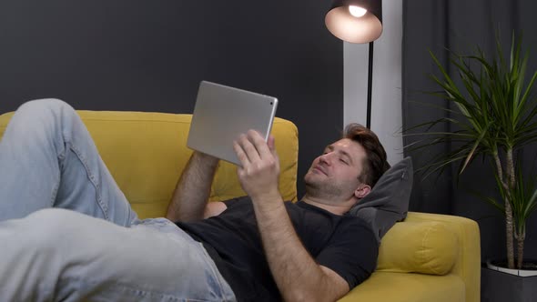 Young Unshaven Handsome Smiling Guy Messaging Online Lying on Sofa with Tablet