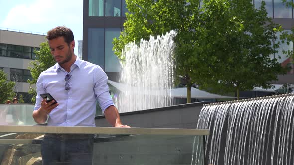 A Young Handsome Man Works on a Smartphone in an Urban Area - Artificial Waterfalls