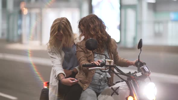 Two Beautiful Young Women Riding a Motorcycle at Night on City Street and Talking to Each Other