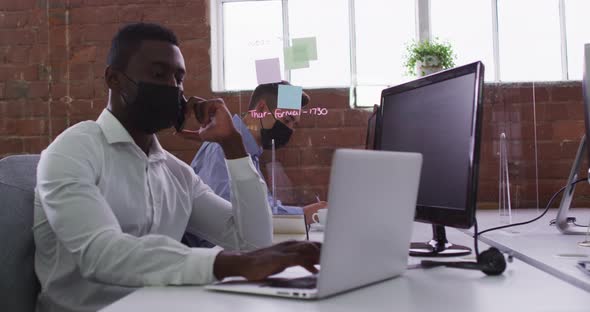 African american businessman wearing mask sitting on desk using laptop talking on smartphone