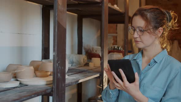 Modern Young Woman Working with Tablet in Pottery Workshop Touching Screen Looking at Ceramics