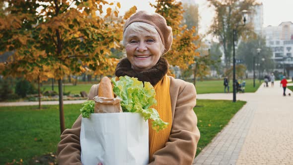 Aged Female is Smiling Holding Paper Bag of Groceries Baguette and Lettuce While Standing in Autumn