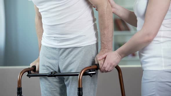 Caring wife helping ill husband to move with walking frame, rehabilitation