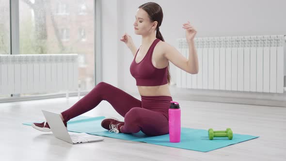 a Woman in a Burgundy Tracksuit with Laptop on a Mat in a Gym