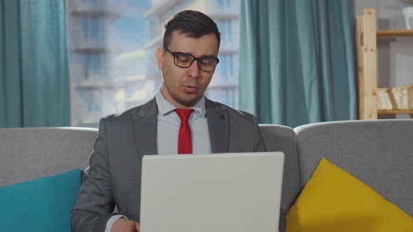 Serious Office Worker in a Suit and Shorts Sits in the Living Room and Talks on a Laptop at Work