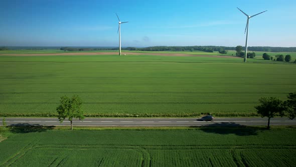 Aerial trucking shot of electric car driving on rural road and spinning wind turbines in background