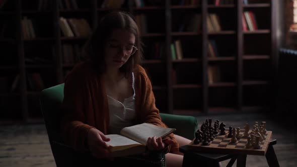 Portrait of Focused Young Intelligent Woman in Glasses Reading Paper Book Sitting at Desk with Chess