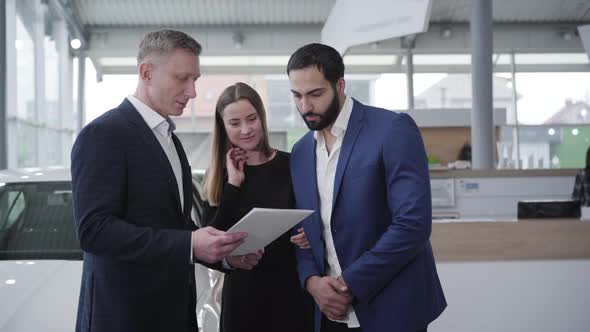 Portrait of Professional Car Dealer Consulting Young Multiracial Family in Showroom. Rich Caucasian