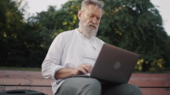 Senior Man with Grey Beard Works on Laptop Sitting on Stairs