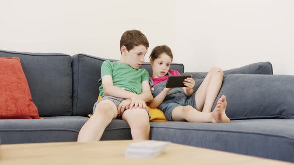 Two kids sitting on a large sofa at home, using a mobile phone