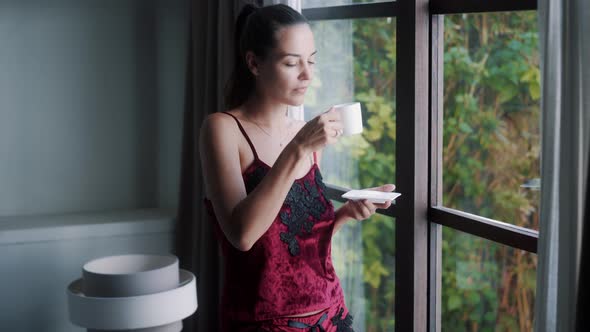 Young Woman in Pajamas Drinking Coffee in Morning and Looking Out Window