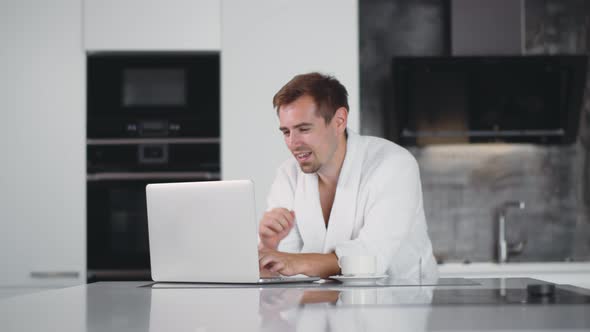Young Handsome Man in Bathrobe Using Laptop and Drinking Coffee at Kitchen in Morning