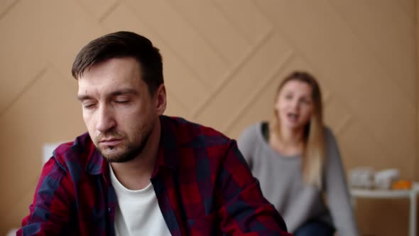 Husband is Sitting on Bed with Straight Face While Wife is Swearing at Him From Behind Front View