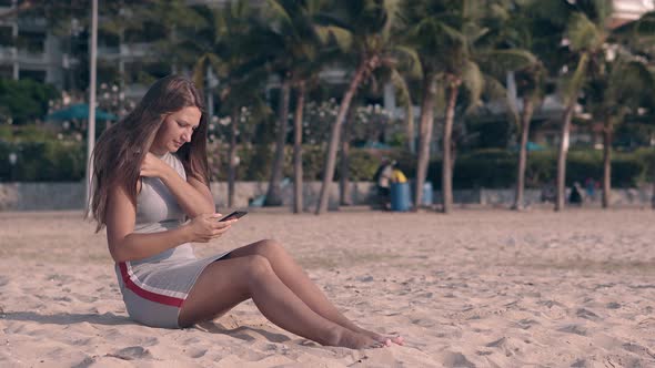 Young Barefoot Woman in Gray Texts on Phone Sitting on Sand