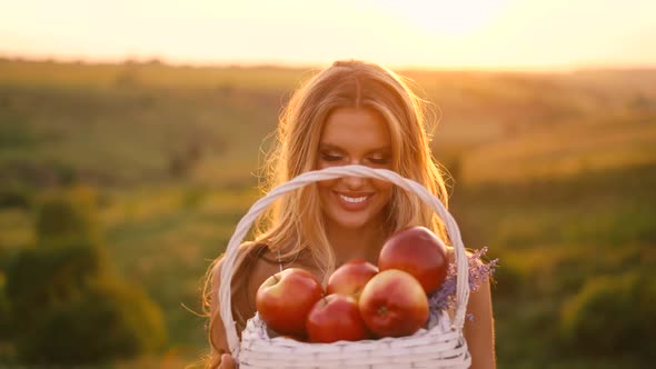 Beautiful sexy blonde girl in white dress posing in a field at sunset with a basket of fruit	