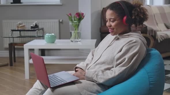 Beautiful Smiling African American Woman in Headphones Sitting on Bag Chair Waving Talking