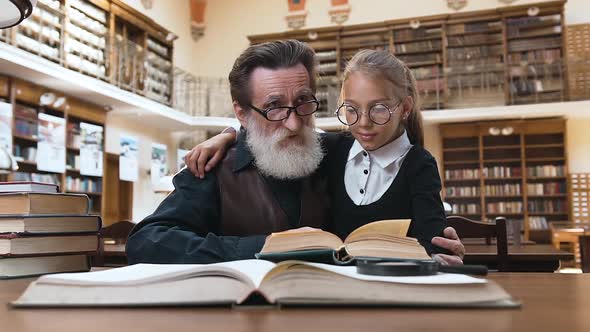 Old Grandpa His Cute Granddaughter which Sitting on Knees while they Reading Book in the Library