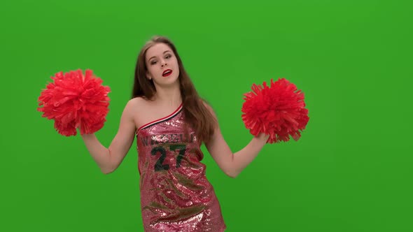 Portrait of a Happy Woman Cheerleader Dancing with Red Pompoms in the Studio on a Green Screen
