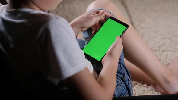 child in shorts on the couch stares into a smartphone with a green screen.