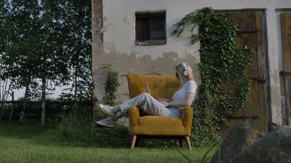 Woman sitting in an armchair using laptop in a garden with wall and doors on background