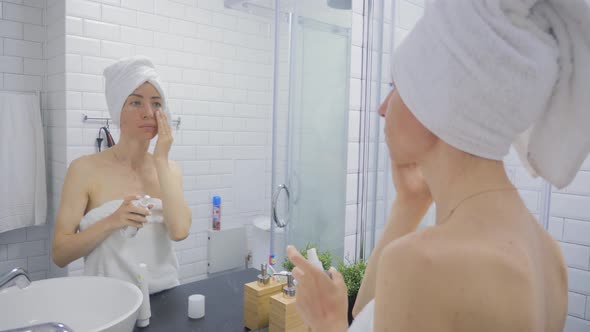 Young Woman in Towel Applying Cream To Face and Looking To Mirror at Home Bathroom. Beauty