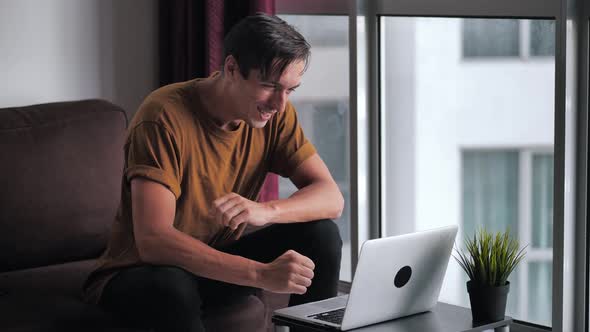 Successful Young Man Using Laptop Gets Surprised Then Happy and Celebrates Win While Sitting on Sofa