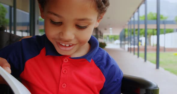 Front view of disabled African American schoolboy reading a book on wheelchair in school corridor 4k