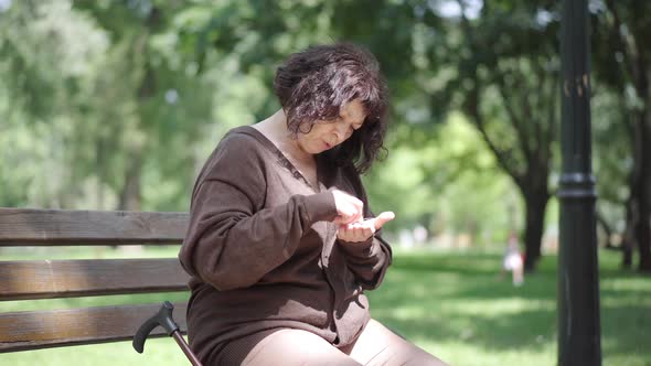 Camera Approaches To Sad Senior Woman Counting Coins on Bench in City Park. Portrait of Unhappy