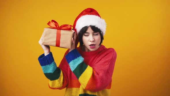 Curious Woman Wearing Santa Hat Shaking Christmas Gift Trying to Guess What's Inside Orange