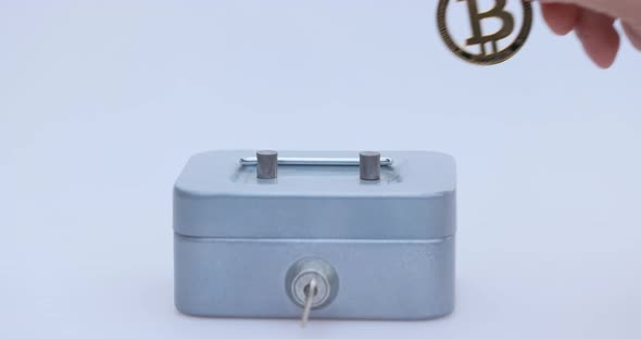 Male Hand Puts a Cryptocoin Bitcoin or BTC in a Small Grey Vault Safe or Piggy Bank Depicting Crypto