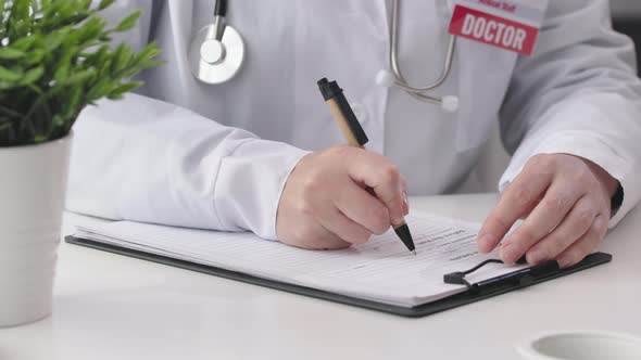 Doctor in White Robe Uniform Red Badge Doctor Sign Sitting Desk Filling Papers