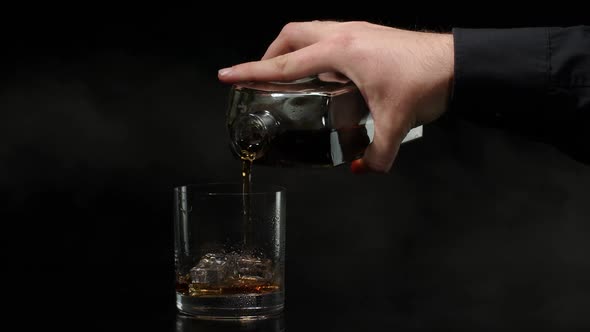 Barman Pouring Whiskey Cognac Brandy From Bottle Into Glass with Ice Cubes on Black Background