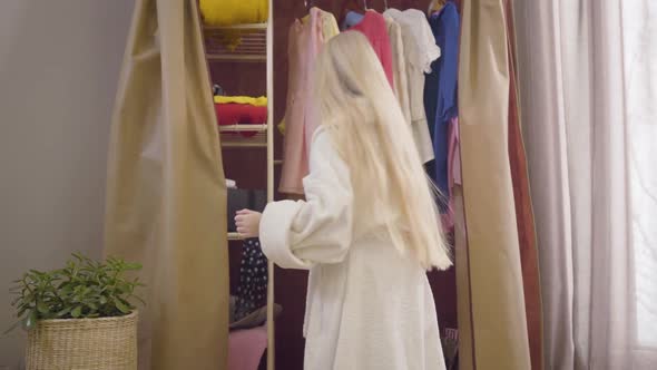 Back View of Little Blond Girl with Long Hair Standing in Front of Open Wardrobe and Looking for