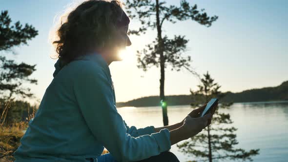 Young Woman Uses Smartphone Sitting on Bank of Tranquil Lake