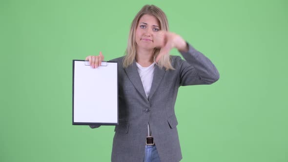 Stressed Young Blonde Businesswoman Showing Clipboard and Giving Thumbs Down