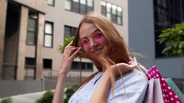 Young Woman in Pink Sunglasses Standing in Front of the Shopping Mall and Holding Shopping Bags