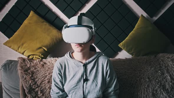 Young Woman Sitting on Sofa Playing Game Using VR Helmet Indoor at Home