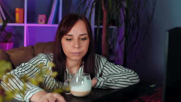 Young Woman Drinks Coffee Sitting in Armchair Before Computer in Office